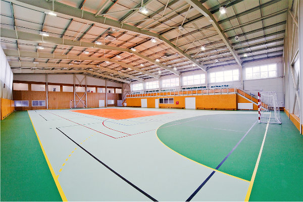 Stahlrahmenkonstruktion der Sporthalle mit Tribüne für Zuschauer im Innenraum und Verbindungskorridor zum Grundschulgebäude.